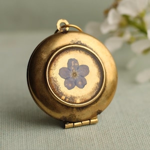 May include: A round gold-toned metal locket featuring a pressed light blue five-petaled flower encased behind a clear circular glass cover. The locket has a small metal loop at the top for attachment and a visible hinge at the bottom edge.