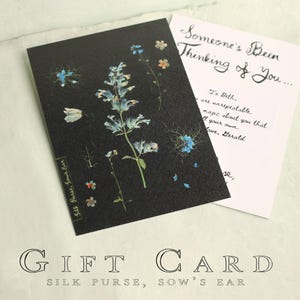 May include: A black gift card with a floral design featuring blue and white flowers. The card is paired with a white card that reads "Someone's Been Thinking of You..." and a handwritten message. The words "GIFT CARD" and "Silk Purse, Sow's Ear" are also visible.