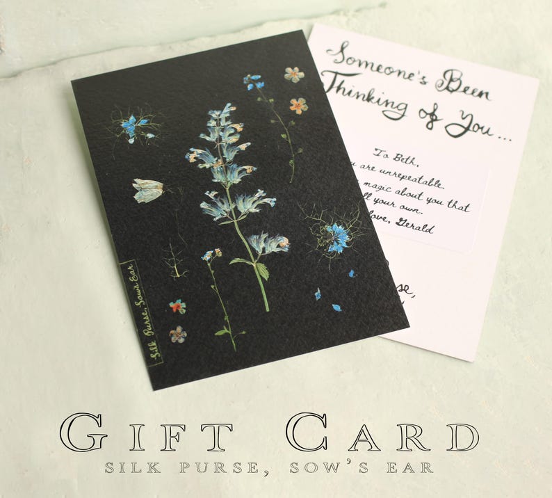 May include: A black gift card with a floral design featuring blue and white flowers. The card is paired with a white card that reads "Someone's Been Thinking of You..." and a handwritten message. The words "GIFT CARD" and "Silk Purse, Sow's Ear" are also visible.