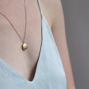 May include: A gold-coloured necklace featuring a delicate chain and a small, smooth, circular pendant. The pendant has a matte finish and is suspended from a simple jump ring, resting against a light blue sleeveless garment with thin straps.