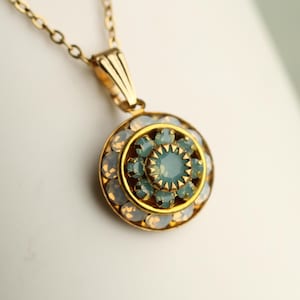 May include: A gold-toned pendant necklace with a circular pendant. The pendant features a central light blue stone surrounded by smaller light blue and opalescent stones. The necklace chain is also gold-toned.
