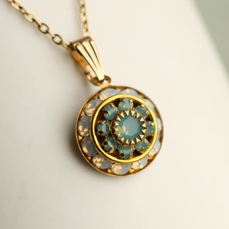 May include: A gold-toned pendant necklace with a circular pendant. The pendant features a central light blue stone surrounded by smaller light blue and opalescent stones. The necklace chain is also gold-toned.