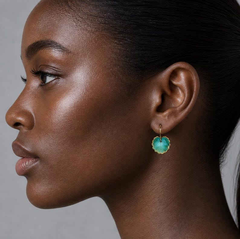 May include: A person's side profile, showcasing a gold earring with a teal, leaf-shaped pendant. The earring is a small, delicate piece of jewellery with a textured edge.