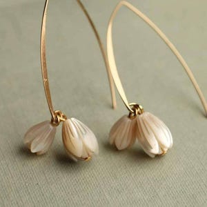 Puede incluir: Un par de delicados pendientes dorados con diseño floral. Cada pendiente tiene un alambre dorado curvado que sostiene dos pequeños colgantes con forma de flor blanco nacarado. Los pendientes están colocados sobre un fondo suave y neutro.