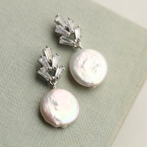 May include: A pair of earrings with a cluster of clear, rectangular crystals above a large, round, lustrous, white pearl. The earrings are set on silver-coloured metal. The pearls have a slightly pinkish hue.