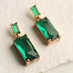 Può includere: Un paio di orecchini dorati con pietre preziose rettangolari verde smeraldo. Ogni orecchino presenta una pietra rettangolare più piccola sopra quella più grande. Gli orecchini sono su uno sfondo bianco.