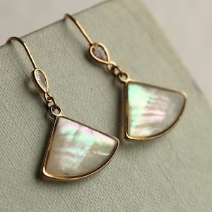 Puede incluir: Un par de pendientes dorados con colgantes iridiscentes en forma de abanico. Los pendientes tienen un diseño delicado, con un pequeño acento en forma de lágrima sobre el abanico. Los colgantes muestran un juego de colores brillantes, incluyendo verde, rosa y blanco.