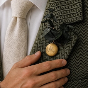 Op de afbeelding: Een rond goudkleurig metalen medaillon met een gegraveerde sierlijke letter M, bevestigd aan een takje met donkergroene eucalyptusbladeren. Het medaillon is opgespeld op de revers van een olijfgroene visgraat blazer over een wit overhemd.