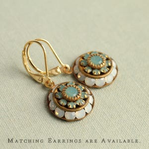 May include: A pair of gold-toned earrings with a round, ornate design. Each earring features a central cluster of light blue stones surrounded by a ring of smaller stones and a border of white, opalescent stones. The text at the bottom reads "Matching Earrings are Available."