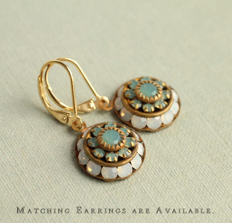 May include: A pair of gold-toned earrings with a round, ornate design. Each earring features a central cluster of light blue stones surrounded by a ring of smaller stones and a border of white, opalescent stones. The text at the bottom reads "Matching Earrings are Available."