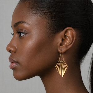 May include: Gold-colored earrings with a fan-like design. The earrings are made of thin, vertical bars that radiate from a central teardrop-shaped element. The earrings are worn on a person's ear.