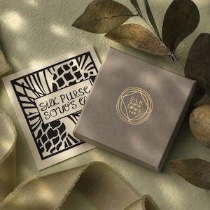 K&ouml;nnte beinhalten: Eine graue quadratische Schachtel und eine Karte mit dem Text "Silk Purse Sows Ear" in Schwarz und Gold. Die Schachtel hat ein goldenes Logo mit dem gleichen Text. Der Hintergrund ist hellgr&uuml;n mit einem Band und Bl&auml;ttern.
