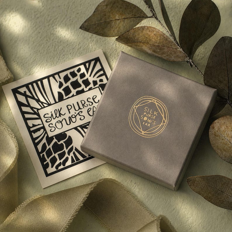 K&ouml;nnte beinhalten: Eine graue quadratische Schachtel und eine Karte mit dem Text "Silk Purse Sows Ear" in Schwarz und Gold. Die Schachtel hat ein goldenes Logo mit dem gleichen Text. Der Hintergrund ist hellgr&uuml;n mit einem Band und Bl&auml;ttern.
