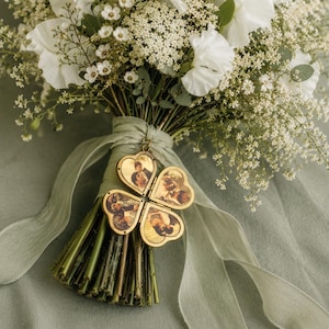 May include: A bouquet of white flowers and greenery tied with a sage green ribbon, featuring a gold-toned, four-leaf clover-shaped locket attached to the stems. The locket is open, revealing four distinct circular photographs of people inside.