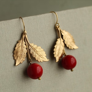May include: Gold-coloured leaf earrings with red cherry-shaped beads. The earrings feature a hook closure and are designed to resemble a branch with leaves and berries. The earrings are a nature-inspired jewellery piece.