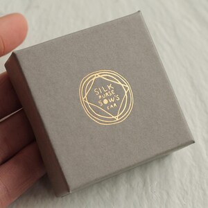 K&ouml;nnte beinhalten: Eine kleine, graue Geschenkbox mit einem goldfarbenen Logo, das "SILK PURSE SOW'S EAR" in einem kreisf&ouml;rmigen Design zeigt. Die Box ist rechteckig und hat eine matte Oberfl&auml;che. Das Logo ist mittig auf der Oberseite der Box angebracht.