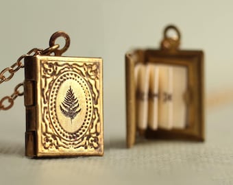 Può includere: Un ciondolo a medaglione in metallo dorato a forma di libro in miniatura, caratterizzato da un design a foglia di felce in rilievo sulla copertina. Il medaglione è fissato a una catenina dorata abbinata e si apre rivelando piccole pagine color crema.