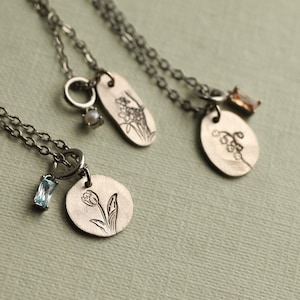 Pode incluir: Três colares de metal prateado com pingentes circulares e ovais gravados com desenhos florais. Cada colar inclui um pequeno berloque de destaque, incluindo uma pérola branca, um cristal retangular azul claro e um cristal castanho.