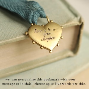 May include: A gold heart-shaped bookmark with the text "here's to a new chapter" engraved on it. The bookmark is attached to a blue ribbon and is resting on a book.