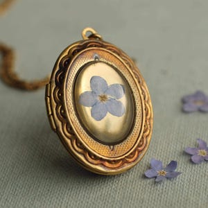 May include: An oval gold-toned metal locket pendant featuring a pressed light blue five-petaled flower encased under a clear domed cover. The locket has a decorative scalloped edge and a textured border, suspended from a matching gold-toned chain.