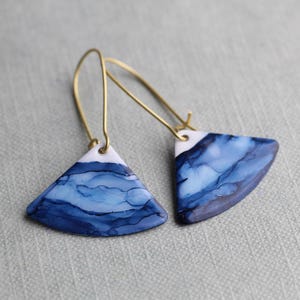 May include: A pair of handmade earrings with a triangular shape. The earrings feature a white top section and a blue, ink-like design. The earrings have gold-coloured ear wires.