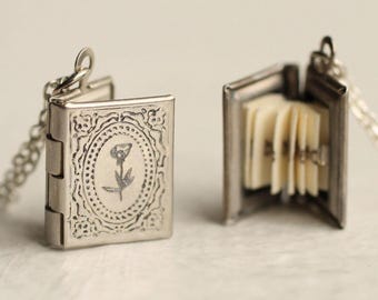May include: A silver-toned metal book locket pendant on a chain. One locket is closed, displaying an embossed oval frame with a central flower engraving. The other locket is open, revealing multiple thin, off-white paper pages inside.