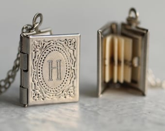 Könnte beinhalten: Ein silberfarbenes Metallmedaillon in Form eines Miniaturbuchs mit aufklappbarem Deckel. Der vordere Deckel zeigt einen geprägten ovalen Rahmen, der den Buchstaben H umschließt. Ein zweites Medaillon ist geöffnet und zeigt mehrere dünne, cremefarbene Papierseiten.