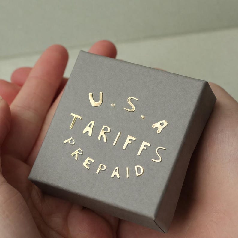 May include: A small, gray box with gold lettering that reads "U.S.A. TARIFFS PREPAID." The box is held in open hands. The box is rectangular and has a simple, clean design.