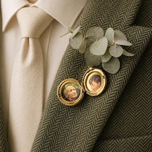 Può includere: Un medaglione d'oro con due piccoli ritratti, uno di una persona anziana e uno di un bambino, è appuntato a una giacca di tweed verde. Un rametto di foglie verdi è infilato sopra il medaglione. Sono visibili una cravatta color crema e una camicia bianca.