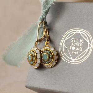 May include: Gold-tone earrings with a round, ornate design featuring a central teal stone surrounded by white and teal accents. The earrings are displayed next to a gray box with the text "SILK PURSE SOW'S EAR" in gold.