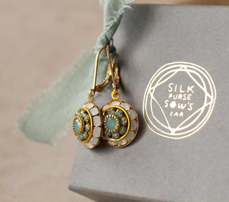 May include: Gold-tone earrings with a round, ornate design featuring a central teal stone surrounded by white and teal accents. The earrings are displayed next to a gray box with the text "SILK PURSE SOW'S EAR" in gold.