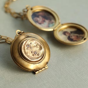 May include: Two gold-toned metal lockets on chains. One locket is closed, featuring a circular center with small star engravings and two embedded clear stones. The second locket is open, revealing two oval-shaped interior frames containing photographs.