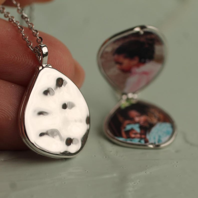Peut inclure: Un collier avec un pendentif en forme de goutte d'eau argent&eacute;, avec une surface &eacute;maill&eacute;e blanche et noire textur&eacute;e. Le m&eacute;daillon est ouvert, r&eacute;v&eacute;lant deux petites photos. Le collier est sur une cha&icirc;ne argent&eacute;e.