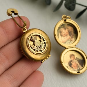 Op de afbeelding: Een ronde koperen medaillon met een uitgesneden distelontwerp aan de voorzijde, bevestigd aan een sluiting in de vorm van een veiligheidsspeld. Daarnaast toont een open koperen medaillon twee ronde vintage-stijl portretten in de vakjes.