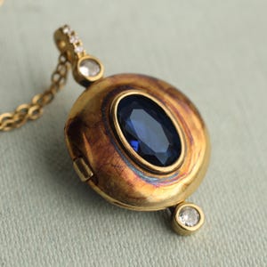 May include: A gold locket pendant featuring a central oval-cut deep blue gemstone. The locket has a textured, weathered gold finish and is accented with small, clear round stones at the top and bottom. It hangs from a delicate gold chain.