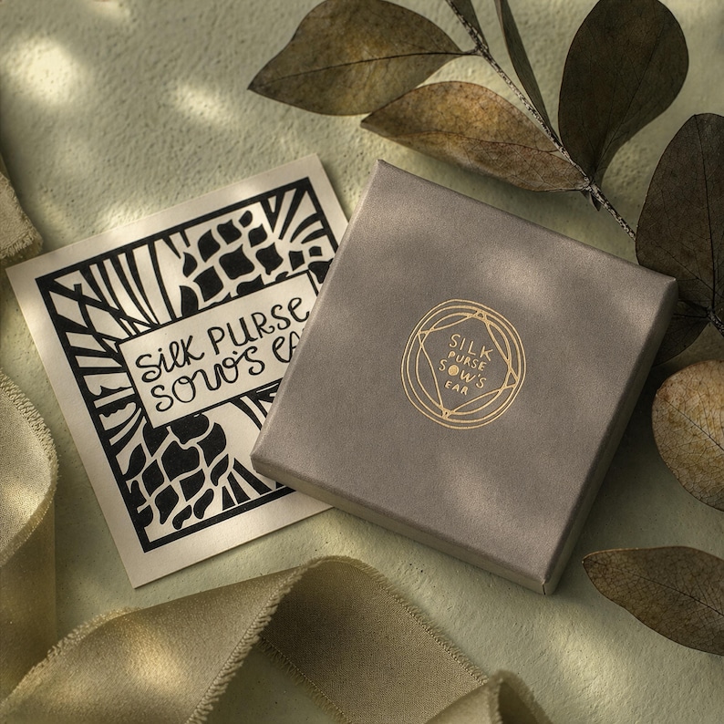 May include: A grey square box and a card with the text "Silk Purse Sows Ear" in black and gold. The box has a gold logo with the same text. The background is a light green with a ribbon and leaves.