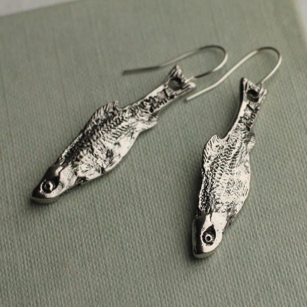 May include: A pair of silver-colored fish-shaped earrings. The earrings are detailed with scales and have a hook closure. The fish are facing opposite directions, and the background is a muted green.