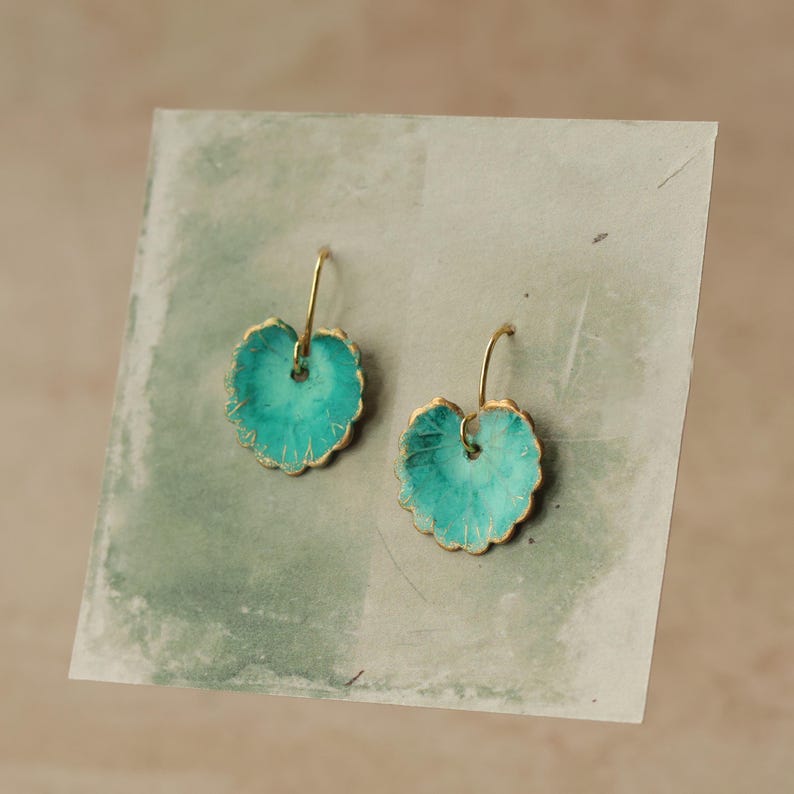 May include: A pair of turquoise leaf-shaped earrings with gold-coloured edges and ear wires. The earrings are displayed on a textured, light green surface. The leaf design has a scalloped edge.