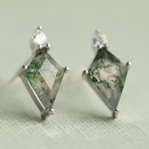 May include: A pair of silver stud earrings. Each earring features a diamond-shaped, moss agate stone with green and white patterns. A small, round, clear stone is set above the diamond shape.