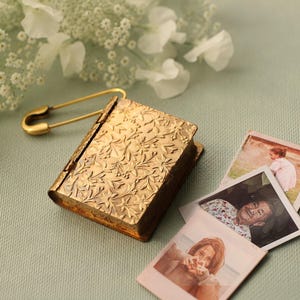 May include: A small, rectangular, gold-toned metal locket shaped like a book with an embossed floral pattern on the cover. A gold safety pin is attached to the spine. Three small, square, white-bordered photographs are fanned out nearby.