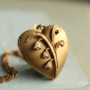 Op de afbeelding: Een goudkleurige hartvormige locket met een bloemmotief. De locket is versierd met drie klokvormige bloemen en een fijne ketting. De locket is gesloten en de details zijn fijn bewerkt.