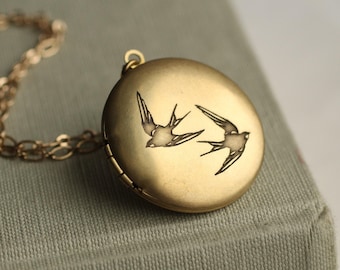 May include: A round, matte gold-toned metal locket pendant featuring an engraved design of two flying swallows. The locket is attached to a matching gold-toned metal chain with oval links, creating a cohesive piece of jewelry with a smooth finish.