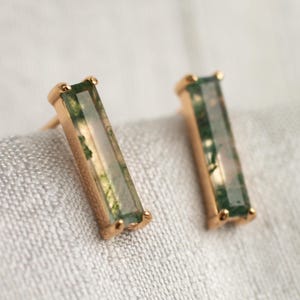 Puede incluir: Un par de pendientes de botón dorados. Cada pendiente presenta una piedra rectangular de ágata musgosa verde engastada en un bisel dorado. Los pendientes se muestran sobre una superficie texturizada de color gris claro.