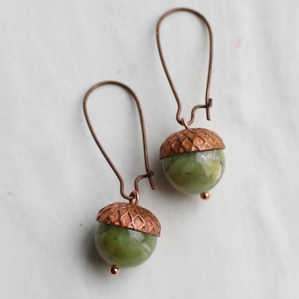 May include: A pair of handcrafted acorn earrings. Each earring features a green, spherical bead with a textured copper-colored cap and a copper-colored ear wire. The earrings are displayed on a white surface.