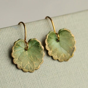 Op de afbeelding: Een paar oorbellen in de vorm van bladeren. Elke oorbel heeft een lichtgroen blad met goudkleurige randen en aderen. De oorbellen hangen aan goudkleurige oorhaken, wat een delicaat, op de natuur geïnspireerd accessoire creëert.