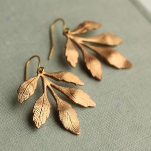 May include: A pair of gold-coloured leaf earrings. Each earring features a cluster of delicate, textured leaves attached to a simple hook. The earrings are displayed on a light blue-grey surface, showcasing their intricate design and warm metallic hue.