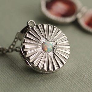 Puede incluir: Un colgante de relicario redondo de plata con un patrón de rayos de sol que irradia desde una piedra de ópalo iridiscente central. El relicario está unido a una delicada cadena de plata, con un segundo relicario abierto visible en el fondo borroso.