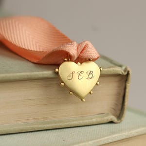 May include: A gold heart-shaped bookmark with the initials "SEB" engraved on it. The bookmark is attached to a peach-colored ribbon.