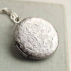 Peut inclure: Un médaillon rond en métal argenté orné d'un motif gravé complexe composé de fleurs et de lignes tourbillonnantes. Le médaillon est fixé à une chaîne en métal argenté, mettant en valeur une surface texturée détaillée sur la face avant.