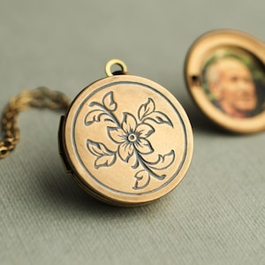 May include: A round, antique-finish brass locket featuring an engraved floral design on the front cover. The locket is attached to a matching brass chain. A second, open locket is visible in the background, displaying a small portrait of a person.